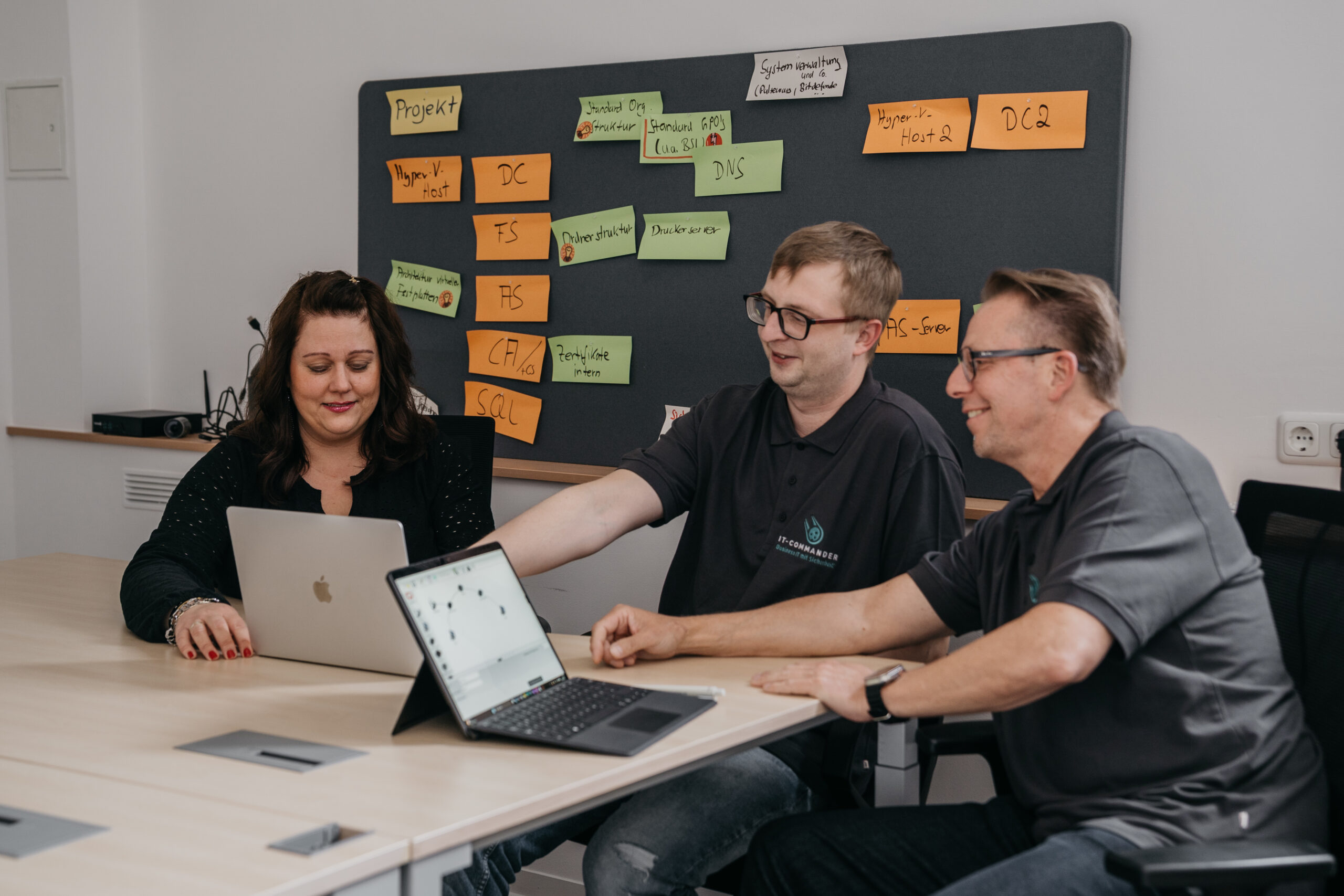 Drei Personen in einem Besprechungsraum, die an Laptops arbeiten und diskutieren.