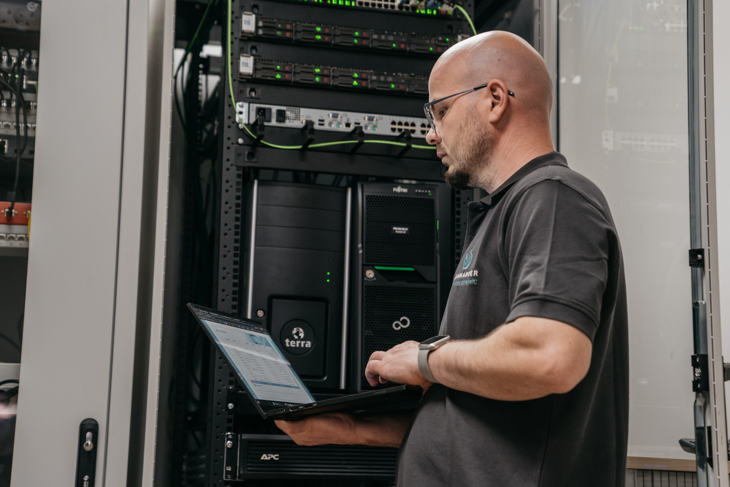 Mann mit Glatze arbeitet an einem Laptop vor einem Serverrack mit mehreren Geräten.